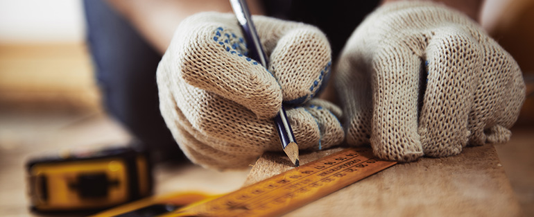 Repairer hands in protective gloves measuring wooden plank with ruler and pencil.